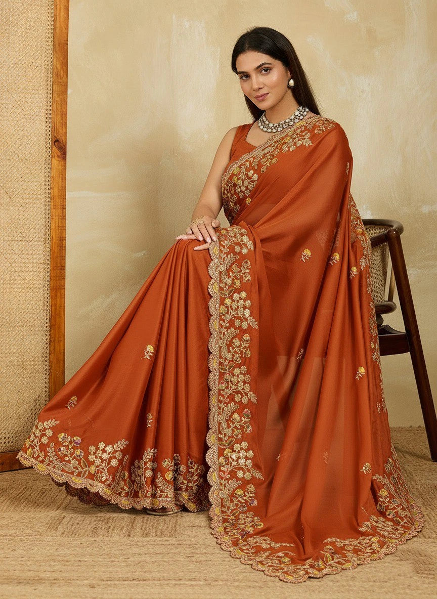 Woman wearing a rust-colored saree with gold embroidery against a beige wall.